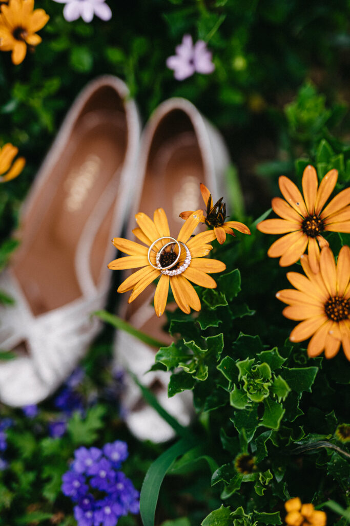 plan semi resserré sur les alliances posée sur un calendula, en arrière plan floutées on voit les chaussure argentées de la mariées entourées d'autre fleurs colorées