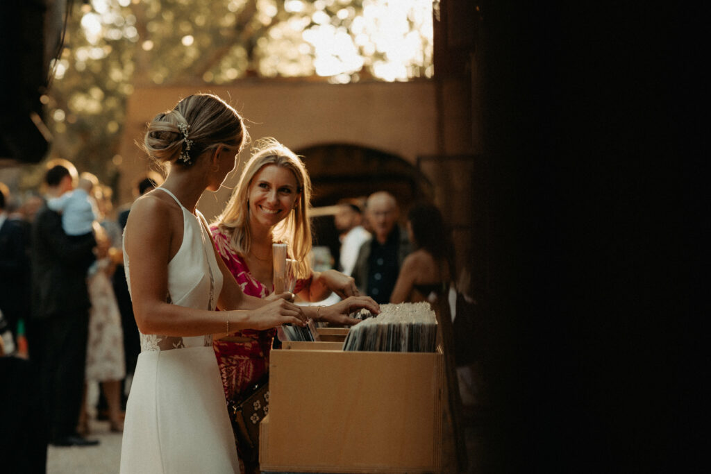plan resserré pris par le photographe mi caché par la pierre de la marriée et une invitée entrain de rire en choisissant de la musique au vin d'honneur, dans la lumière chaleureuse de fin d'après midi.